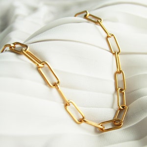 May include: A gold chain necklace with rectangular links. The chain is on a white fabric background.