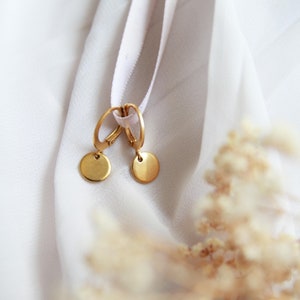 May include: A pair of gold hoop earrings with small round discs hanging from them. The earrings are on a white fabric background.