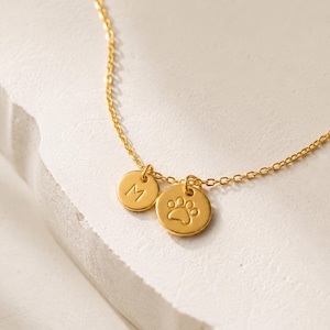 May include: A gold-toned chain necklace featuring two small circular charms. One charm is engraved with the letter M, and the other charm features an embossed paw print design. The delicate metal chain connects to both circular pendants.