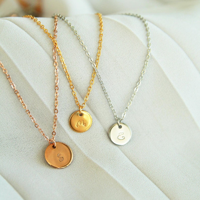 Puede incluir: Tres collares de oro con colgantes de iniciales. Los collares est&aacute;n sobre una superficie blanca texturizada. Las iniciales son M, S y E.