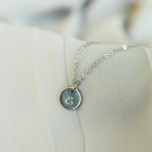 Puede incluir: Un collar de cadena de plata con un peque&ntilde;o colgante redondo con la letra "E" grabada.