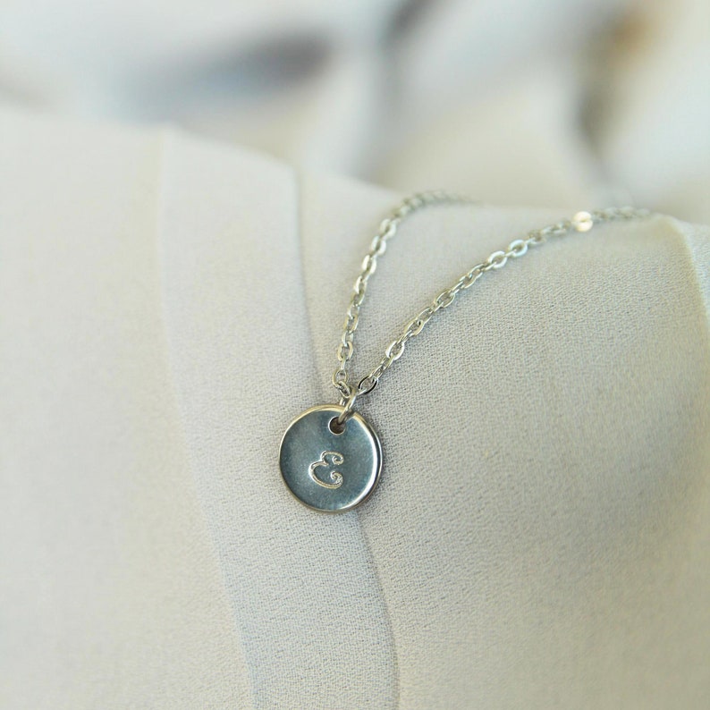 Puede incluir: Un collar de cadena de plata con un peque&ntilde;o colgante redondo con la letra "E" grabada.
