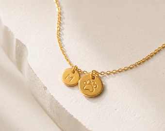 May include: A gold-coloured chain necklace featuring two small, round, flat charms. One charm is engraved with the capital letter M, and the other charm is engraved with a paw print design. The necklace is made of thin, delicate metal links.