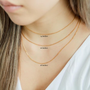 Puede incluir: Tres cadenas de oro de diferentes longitudes, 14 pulgadas, 16 pulgadas y 18 pulgadas, se muestran en el cuello de una modelo.