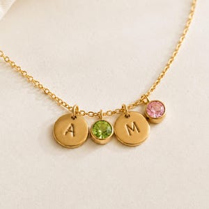 May include: A gold-toned chain necklace featuring two circular charms engraved with the letters A and M, accompanied by two round bezel-set gemstones in light green and light pink, all suspended from a delicate link chain.