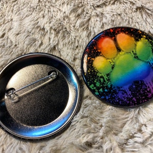 May include: A rainbow paw print on a black background with white splatters. The back of the button shows a silver pin.
