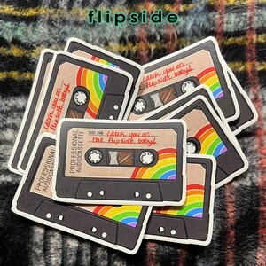 May include: A stack of cassette tape stickers with a rainbow design and the text "catch you on the flipside, baby!" printed on them. The stickers are designed to look like vintage cassette tapes.