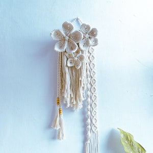 May include: A cream macrame wall hanging with three flower shapes and a butterfly design. It features knotted details, tassels, and wooden beads. The wall decor is displayed against a light blue wall, creating a bohemian style.
