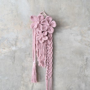 May include: A pink macrame wall hanging with a floral design. The hanging features a variety of knots and tassels.