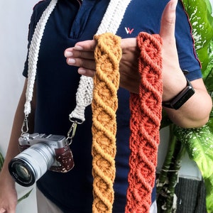 Puede incluir: Tres correas para cámara hechas de cordón de macramé. Las correas son de color blanco, amarillo mostaza y naranja quemado. Las correas se muestran sostenidas por una persona.