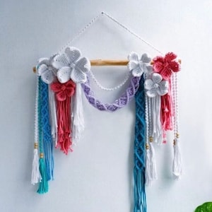 May include: A white wall hanging with a wooden dowel and colorful macrame flowers. The flowers are made with white, pink, and red yarn. The macrame is made with white, pink, blue, and purple yarn. The wall hanging has tassels and beads.