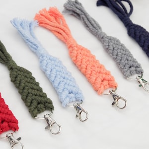 May include: A collection of colorful macrame keychains in various shades, including red, green, blue, peach, gray, and navy. Each keychain features a woven design and a silver-colored clasp for attaching keys or accessories.