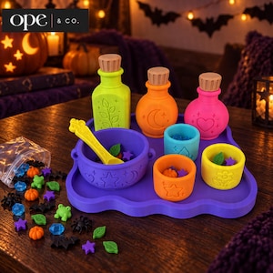 May include: A purple silicone tray holding a yellow spoon, a purple cauldron, three small cups in blue, orange, and yellow, and three bottles in lime green, orange, and pink. The set includes small loose pieces like frogs, pumpkins, and stars.