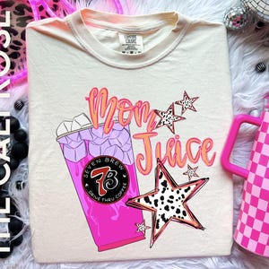 May include: Cream-colored t-shirt with the words "Mom Juice" in orange and pink script, along with a graphic of a pink drink and dalmatian print stars. The shirt also features the text "Seven Brew Drive Thru Coffee".