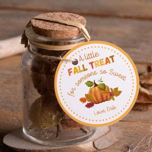 Może przedstawiać: Przezroczysty szklany słoik z korkową pokrywką i ozdobną etykietą. Etykieta głosi "A little FALL TREAT for someone so Sweet" z ilustracją dyni i jesiennych liści. Słoik stoi na drewnianym spodku.
