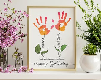 May include: A framed print featuring two handprints in orange and red watercolor. The names "Emma" and "Victoria" are written below the handprints. The text "Happy Birthday 2023" is at the bottom, with the message "Thank you for helping us grow, Mommy!"