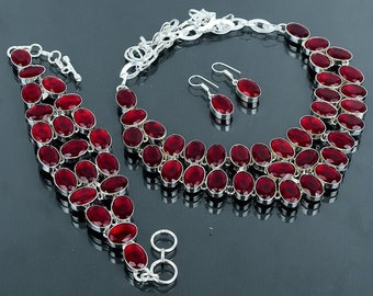 以下が含まれることがあります： 赤い宝石のアクセントが付いたシルバーのネックレス、ブレスレット、イヤリングのセット。ネックレスとブレスレットは、複数の列の楕円形の赤い石が特徴です。