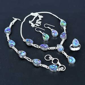 May include: A silver jewellery set comprising a necklace, bracelet, earrings, and ring. Each item features a teardrop-shaped abalone shell in blue, green, and purple hues. The set is presented on a black surface.