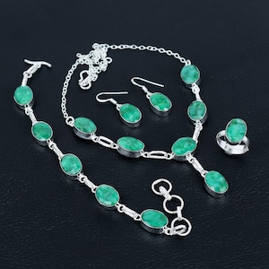 Può includere: Un set di gioielli completo con collana, bracciale, orecchini e anello. Ogni pezzo ha una montatura argentata ed è adornato con pietre preziose verdi ovali. Il set è esposto su una superficie scura.
