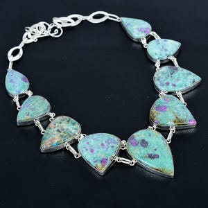 May include: A silver necklace with turquoise, teardrop-shaped stones, speckled with purple and brown. The stones are linked together with silver chain. The necklace is presented on a black backdrop.