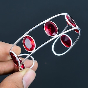 Può includere: Un braccialetto rigido in argento con cinque pietre ovali rosse. Il braccialetto è aperto e ha un design semplice.