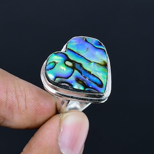 Puede incluir: Anillo en forma de corazón con una pieza central de concha de abulón iridiscente. La concha muestra tonos de azul, verde y morado, engastada en una banda de plata. Fondo oscuro.