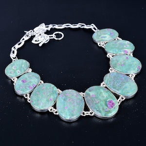 May include: A necklace featuring oval-shaped green and pink gemstones set in silver. The gemstones are connected by a silver chain. The necklace is displayed against a black background.