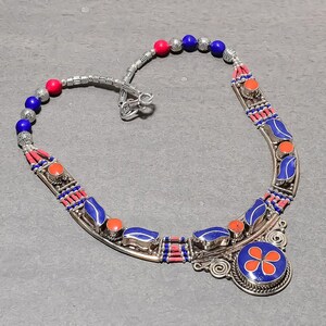 May include: A silver-toned necklace with red, blue, and orange accents. The necklace features a floral pendant and a series of blue and orange stones. The necklace is made of metal and beads.