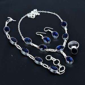 May include: A complete jewelry set featuring a necklace, bracelet, earrings, and ring. Each piece has a silver-tone metal setting with oval, deep blue gemstones. The set is displayed on a black surface.