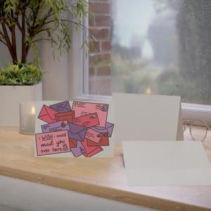 May include: Greeting card with a pile of illustrated envelopes in shades of pink, red, and purple. The card has the text "I wish I could mail you over here." Two blank white cards are also present.