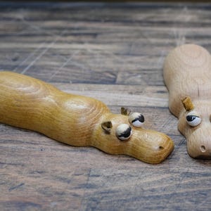 Puede incluir: Dos figuras de hipopótamos de madera. Uno está tumbado, el otro de pie. Ambos tienen ojos grandes y expresivos y orejas pequeñas y marrones. La madera tiene un tono cálido y natural, y los detalles están cuidadosamente tallados.