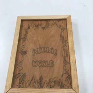Pode incluir: Placa de madeira com as palavras "ANIMAL WORLD" em uma fonte l&uacute;dica. A placa &eacute; emoldurada com madeira clara e apresenta uma borda gravada detalhada de folhas e folhagens tropicais. O fundo &eacute; de cor marrom claro.