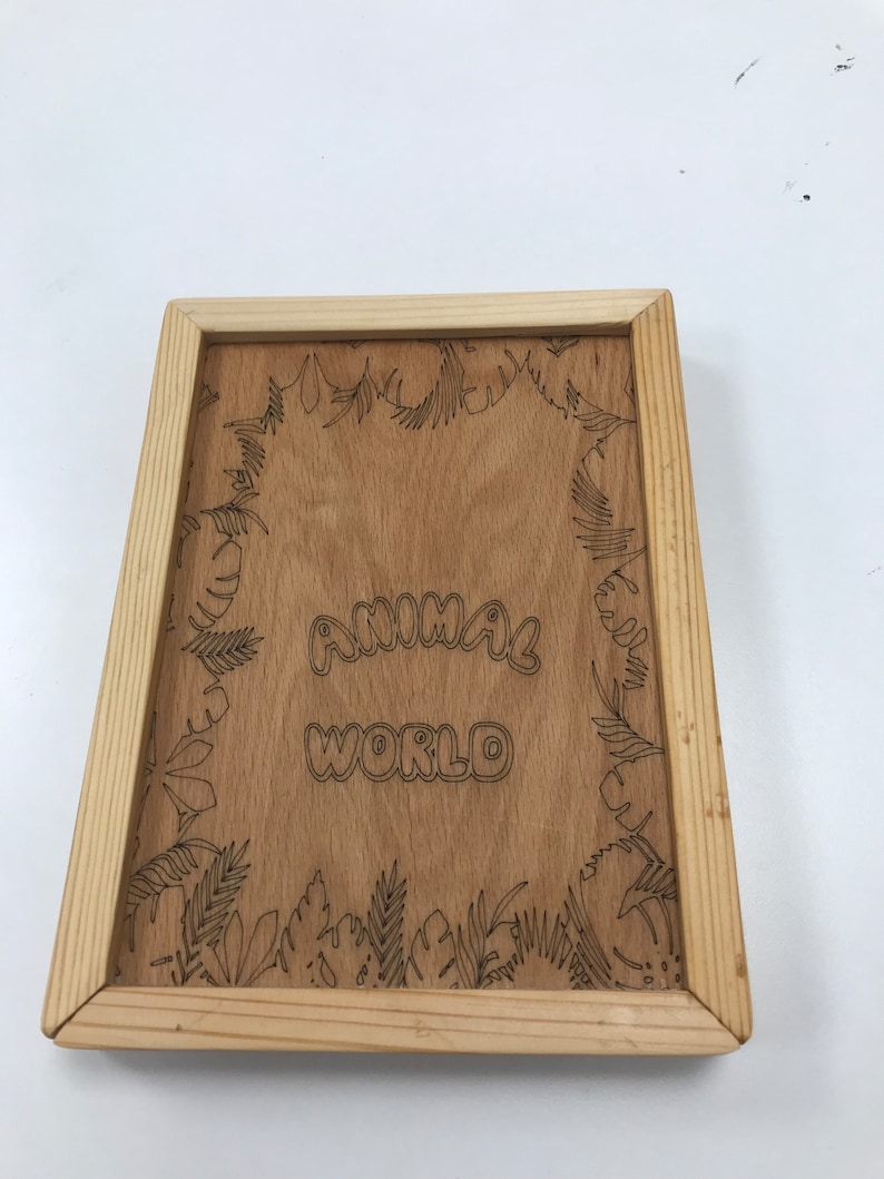 Pode incluir: Placa de madeira com as palavras "ANIMAL WORLD" em uma fonte l&uacute;dica. A placa &eacute; emoldurada com madeira clara e apresenta uma borda gravada detalhada de folhas e folhagens tropicais. O fundo &eacute; de cor marrom claro.