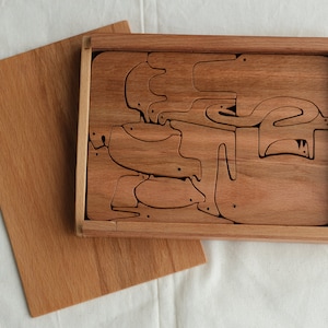 Op de afbeelding: Een houten puzzel met dierenfiguren, waaronder vogels en vissen, wordt gepresenteerd in een rechthoekige houten lijst. De puzzelstukjes zijn lichtbruin. Een apart houten bord ligt naast de lijst. De achtergrond is een witte doek.