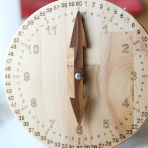 May include: A wooden clock with a light-coloured face and dark brown hands shaped like arrows. The numbers 1 to 12 are carved into the wood, with smaller numbers marking the minutes. The clock is a decorative item.