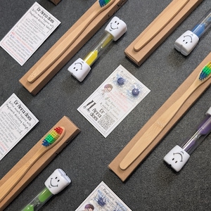 May include: A selection of bamboo toothbrushes with colourful bristles, presented in cardboard packaging. The image also shows smiley-faced timers and small cards with instructions. The items are arranged on a dark surface.