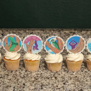 May include: Six cupcakes with white icing, each decorated with a circular image of a cartoon monkey or bird. The images depict scenes with trees, balloons, and a sunset. The cupcakes are arranged on a speckled countertop.