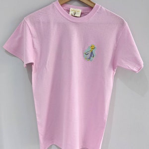 May include: A pink t-shirt with a cartoon character embroidered on the front. The character is yellow with blue and green clothing. The text "Little Tape" is on the tag.