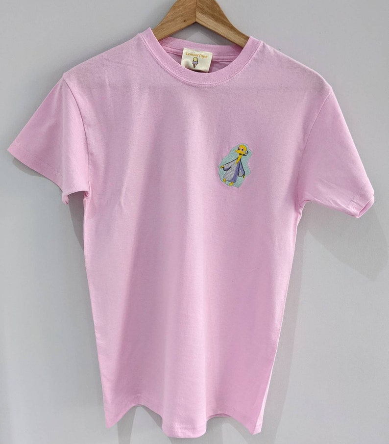 May include: A pink t-shirt with a cartoon character embroidered on the front. The character is yellow with blue and green clothing. The text "Little Tape" is on the tag.