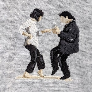 May include: Embroidered image of two figures in black and white, dancing. The figure on the left is wearing a white shirt and black pants, while the figure on the right is wearing a black suit.