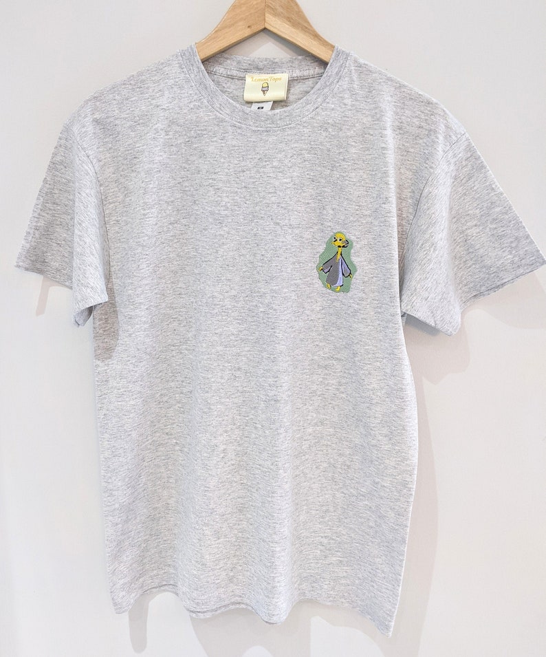 May include: A heather gray t-shirt with a cartoon illustration of a person wearing a purple robe and a yellow crown. The text "Lemon Drop" is on the tag.