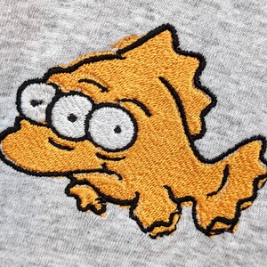 May include: Embroidered yellow fish with three eyes and a black outline on a gray background.