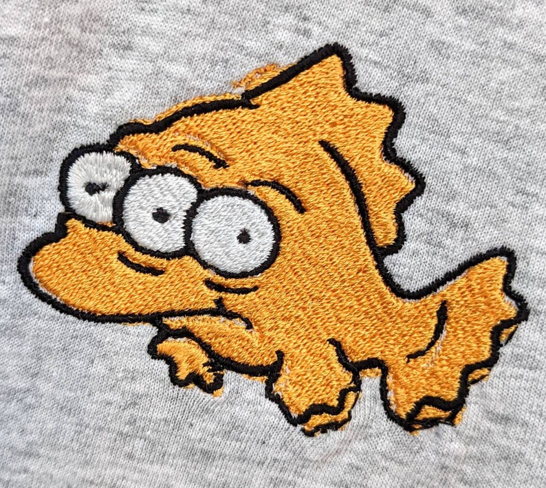 May include: Embroidered yellow fish with three eyes and a black outline on a gray background.