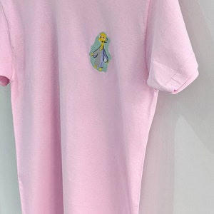 May include: A pink t-shirt with a cartoon character embroidered on the front. The character is yellow with a blue and green outfit. The text "Lemon Tops" is on a tag attached to the shirt.
