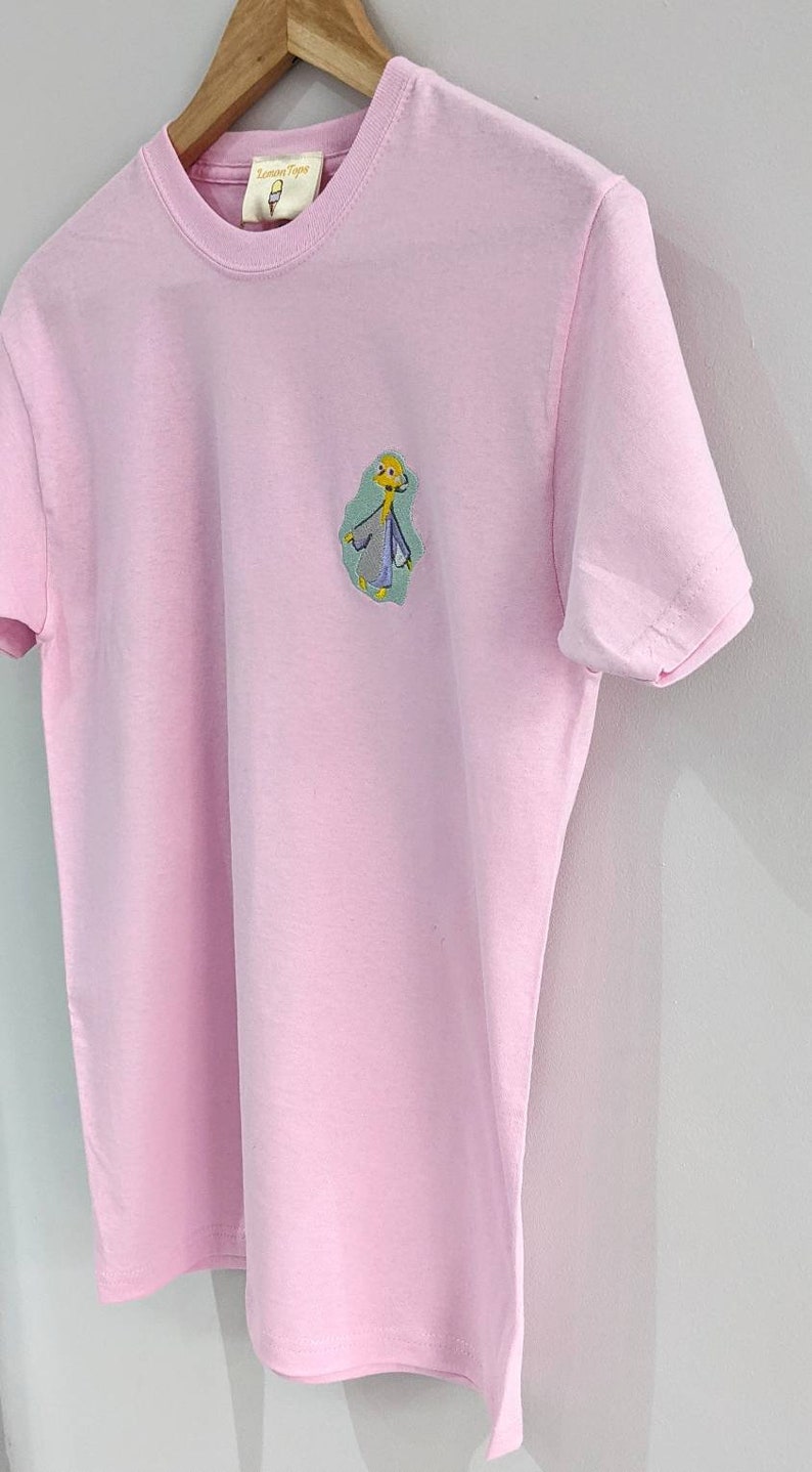 May include: A pink t-shirt with a cartoon character embroidered on the front. The character is yellow with a blue and green outfit. The text "Lemon Tops" is on a tag attached to the shirt.