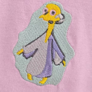 May include: Embroidered patch of a cartoon character wearing a purple and gray robe. The character has yellow skin, black eyes, and a yellow beak. The background is light blue.