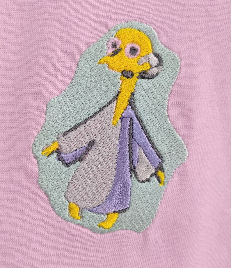 May include: Embroidered patch of a cartoon character wearing a purple and gray robe. The character has yellow skin, black eyes, and a yellow beak. The background is light blue.