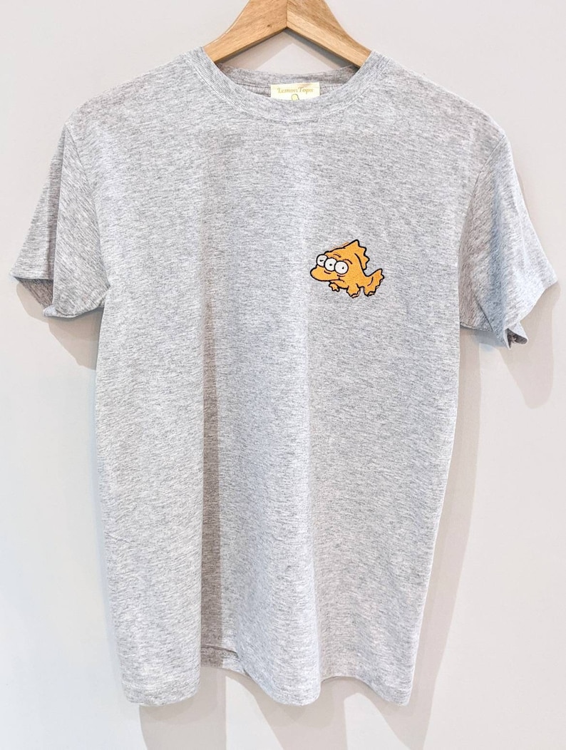 May include: A heather grey t-shirt with a cartoon image of a yellow character with three eyes. The character is wearing a blue shirt and has a yellow fin on its back.