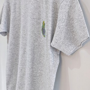 May include: A heather grey t-shirt with a cartoon character graphic on the left chest. The character is green, yellow, and blue.