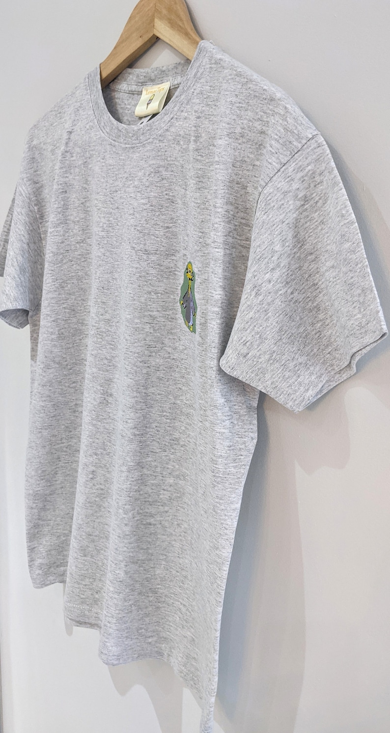 May include: A heather grey t-shirt with a cartoon character graphic on the left chest. The character is green, yellow, and blue.